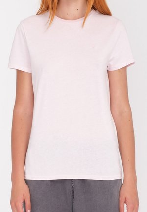 Kobieta ubrana w prosty jasnoróżowy t-shirt z krótkim rękawem i szare spodnie, stoi na jednolitym białym tle.