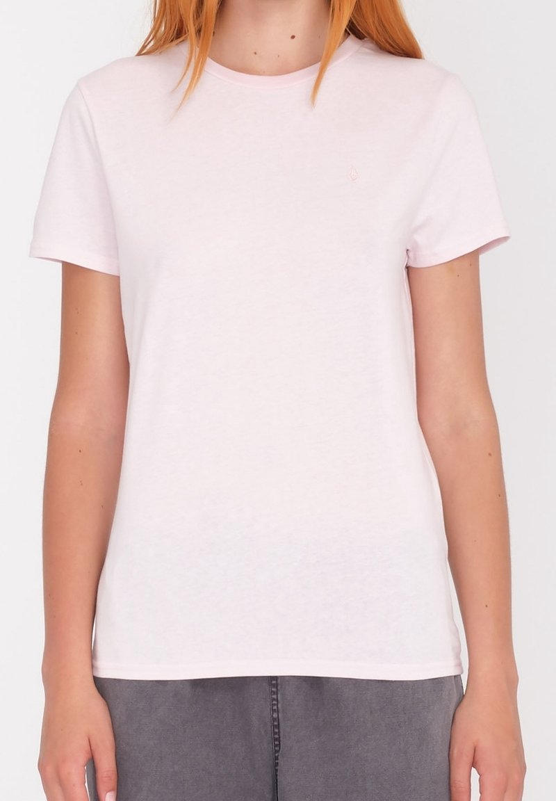 Femme portant un t-shirt rose clair uni à manches courtes avec un pantalon gris, debout devant un fond blanc uni.