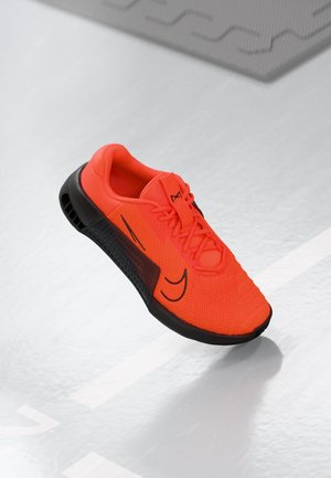 Chaussure de sport orange vif avec semelle noire et logo swoosh noir, flottant au-dessus d'un sol gris clair avec des marquages blancs.