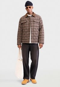 Jachetă matlasată în carouri în nuanțe de maro, cămașă bej, pantaloni negri, cizme galbene și o geantă de umăr albă. Accesoriile includ o căciulă tricotată și ochelari de soare.