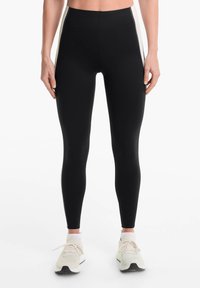 Leggings noirs taille haute avec une texture lisse et des rayures blanches sur les côtés, associés à des chaussures de sport de couleur claire.
