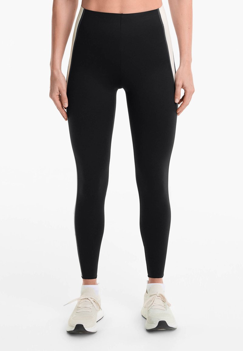 Leggings noirs taille haute avec une texture lisse et des rayures blanches sur les côtés, associés à des chaussures de sport de couleur claire.