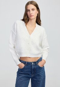 Kobieta w białym, krótkim kardiganie zapinanym na guziki z drobnymi plamkami oraz wysokim stanie niebieskich dżinsów, z rękami w kieszeniach, o neutralnym wyrazie twarzy.