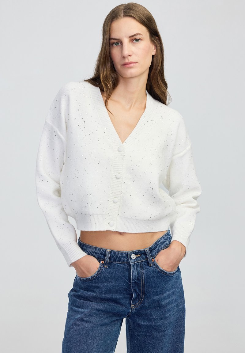 Kobieta w białym, krótkim kardiganie zapinanym na guziki z drobnymi plamkami oraz wysokim stanie niebieskich dżinsów, z rękami w kieszeniach, o neutralnym wyrazie twarzy.