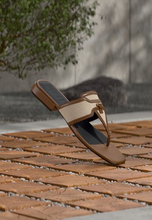 Sandale tip tanga maro și bej cu talpă căptușită plutind deasupra unui podea de placă din lemn cu vegetație și cu un perete de beton pe fundal.