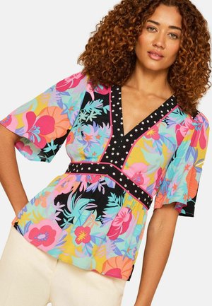 Kobieta o kręconych włosach ubrana w kolorową kwiatową bluzkę z bufkami, wykończoną czarno-białym groszkowym akcentem, oraz kremowe spodnie.