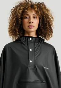 Svart vattentät poncho med hög krage, framsnappar, justerbar huva och framficka. Slät struktur med matt finish.