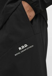 Melnas auduma džemperbikses ar sānu kabatu un baltu uzdrukātu tekstu "R.D.D. ROYAL DENIM DIVISION". Gluda tekstūra un brīvi pieguļoša piegriezums.