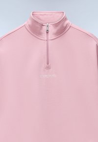 Ανοιχτό ροζ φούτερ με φερμουάρ μέχρι τη μέση, με ανεβασμένο γιακά και λευκή κεντητή επιγραφή "NAPAPIJRI" στο κέντρο κάτω από το φερμουάρ.