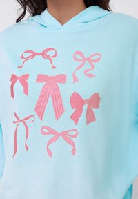 Felpa azzurra con cappuccio decorata con otto disegni di fiocchi rosa in vari stili e dimensioni, con un mix di motivi a tinta unita e a righe.