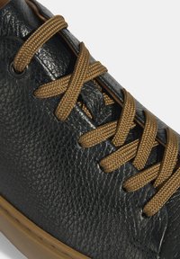 Sneaker en cuir noir présentant une surface texturée, des lacets marron et une semelle en caoutchouc, mettant en avant un design global élégant et moderne.