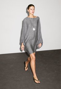 Abito in lino grigio con maniche lunghe e voluminose, di lunghezza al ginocchio e vestibilità comoda. Abbinato a infradito nere e a una collana con pendente in argento.