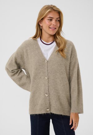 Jeune femme souriante portant un cardigan beige duveteux boutonné, une chemise blanche, un jean foncé et un collier de perles violet sur un fond clair uni.