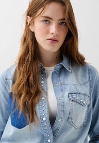 Lichtblauwe denim shirt met een knoopsluiting aan de voorkant, één borstzak en een donkerblauwe patch. Geprofileerde stof en zilveren knoopaccenten.