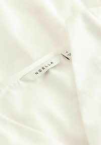 Λευκό ύφασμα με ραμμένη ετικέτα που γράφει «NOELLA» και μια μικρότερη ετικέτα με το μέγεθος «S» και την ένδειξη «Κατασκευάστηκε στην Κίνα».