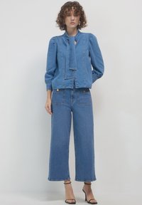 Giacca di denim con collo a cravatta, maniche a sbuffo e bottoni sul davanti. Abbinata a jeans a gamba larga, entrambi in un lavaggio azzurro chiaro. Tacchi neri con lacci.