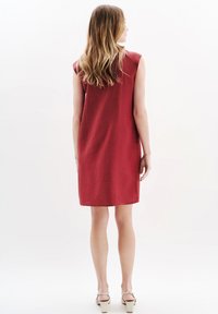 Punainen hihaton mekko, jossa on rento istuvuus, polvipituinen, kankaassa on hienovarainen rakenne, yhdistetty vaaleissa korkokenkissä.