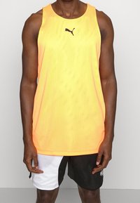Hombre vistiendo una camiseta deportiva sin mangas amarilla de Puma y pantalones cortos de baloncesto en blanco y negro de Puma, de pie frente a un fondo liso.