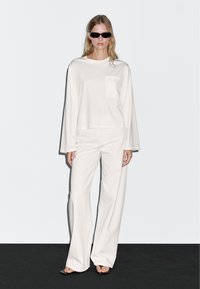 Weißes Langarmhemd mit einer Brusttasche, kombiniert mit weißen weit geschnittenen Hosen. Das Model trägt schwarze Sonnenbrillen und sandalen.