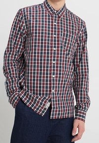 Chemise à carreaux à boutons avec un motif en grille rouge, bleu et blanc. Manches longues, col, poche avant et boutons blancs sur un tissu léger.