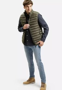 Lekką oliwkowo-zieloną kamizelkę puchową noszoną na ciemnoniebieskiej koszuli, w połączeniu z jasnoniebieskimi dżinsami i stonowanymi brązowymi butami na sznurówki.