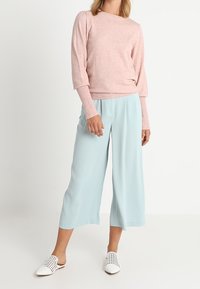 Žena v světle růžovém dlouhém rukávu, světle modrých širokých kalhotách po kotníky a bílých tkaných slip-on botách, stojící na bílém pozadí.