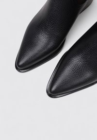 Zapatos de piel de cuero negros con punta afilada, acabado texturizado, silueta estilizada y bordes suaves. Presentan una suela brillante y un diseño minimalista.