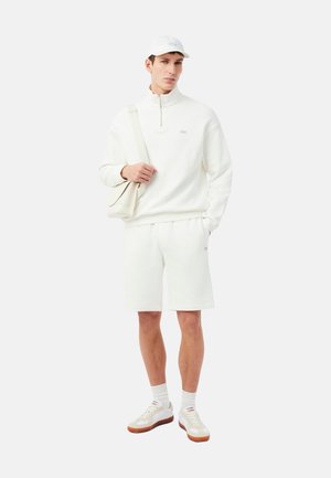 Hombre con sudadera blanca de cremallera hasta el pecho, pantalones cortos a juego, gorra blanca, bolso crema colgado del hombro y zapatillas blancas con suelas de goma.