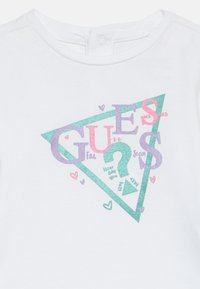 Weißes Shirt mit buntem glitzerndem "GUESS"-Text in einem türkisfarbenen Dreieck, verziert mit kleinen Herzen und einem Fragezeichen.