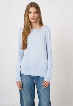 Młoda kobieta o długich, prostych włosach, ubrana w jasnoniebieski sweter z długim rękawem i luźne niebieskie dżinsy, stoi na tle jednolitego tła.