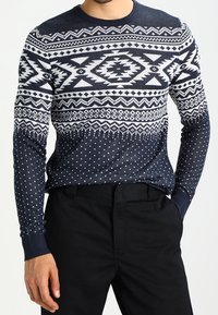 Mörkblå stickad tröja med vita geometriska mönster, inklusive diamanter och prickar, samt ribbade ärmar. I kombination med svarta byxor.