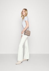 Camiseta lila, pantalones acampanados blancos y bolso cruzado beige con una textura suave. Modelo con botas de tobillo beige con un patrón sutil.