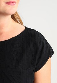 Zwarte textuurtop met een ruime hals en korte mouwen, met een patroon in een gebreide stof. Gladde stof met een subtiele glans.