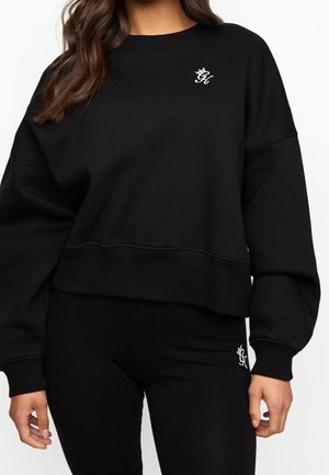 Kvinde iført sort mellemlang cropped sweatshirt med lange ærmer og matchende leggings, begge med lille hvid kursiv "GK"-logo på bryst og lår.