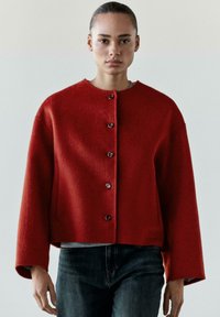 Giacca di lana rossa con scollo rotondo, bottoni frontali e maniche larghe. Tessuto liscio e lunghezza corta, abbinata a jeans scuri.