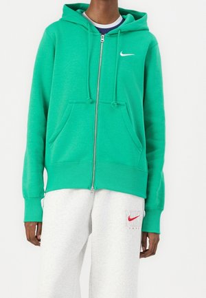 Osoba ubrana w zieloną bluzę z kapturem zapinaną na zamek błyskawiczny z białym logo Nike w kształcie łyżwy oraz białe spodnie dresowe z czerwonym logo Nike i napisem na lewej udzie.