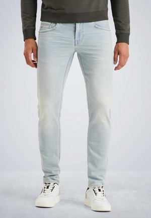 Mann trägt hellblaue Slim-Fit-Jeans und weiße Turnschuhe, steht vor einem schlichten Hintergrund.