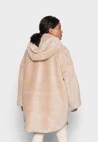 Manteau en polaire beige et moelleux avec capuche, design surdimensionné et fentes latérales. Tissu texturé, manches longues, tombe sous les hanches.
