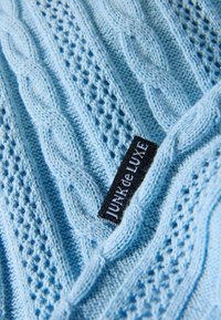 Maglione azzurro chiaro lavorato a maglia con motivi a trecce e trafori, con un'etichetta nera visibile con la scritta "JUNK de LUXE".