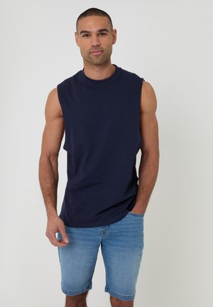 Mand iført navy ærmeløs skjorte og lyseblå cowboyshorts står mod hvid baggrund.