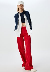 Rote weit geschnittene Hosen mit glatter Textur, kombiniert mit einer marineblauen Zip-Jacke über einem schwarzen Crop-Top. Dazu gehört eine leichte Mütze und weiße Schuhe.