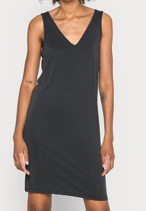 Czarna sukienka bez rękawów z dekoltem w serek, sięgająca do kolan, noszona przez osobę z dwoma widocznymi tatuażami na ramionach, stojącą na jednolitym tle.