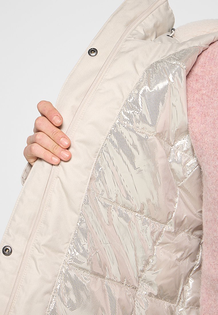 Hand, die einen offenen beigen Mantel hält, der mit einer glänzenden, steppartigen Isolierung gefüttert ist, die kleine Perforationen aufweist, und einen rosa Fleece-Ärmel sichtbar hat.