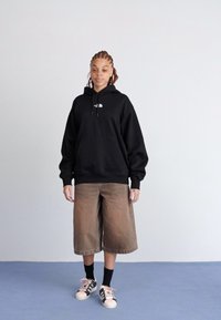 Haut à capuche noir avec logo blanc, shorts larges beiges, et baskets accentuées de rose. Tissu doux, coupe décontractée, avec une poche avant sur le haut à capuche.