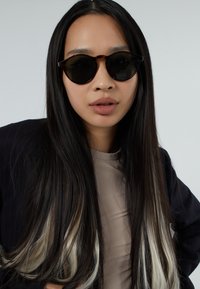 Zonnebril met donkere glazen in een zwart schildpadkleurig montuur. De drager heeft lang, steil zwart haar met blonde highlights, en draagt een beige top en een zwarte blazer.