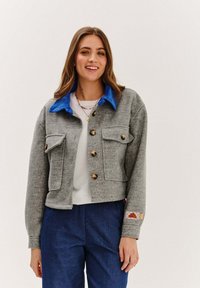 Mladá žena sa usmieva, oblečená v šedej preklápacej bunde s modrým golierom, bielej blúzke, modrých nohaviciach a s viacerými náhrdelníkmi, stojaca na jednoduchom pozadí.