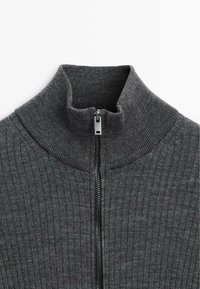 Grauer, gerippter Pullover mit hohem Kragen und silbernen Reißverschlussdetails vorne, der ein weiches, strukturiertes Material zeigt.
