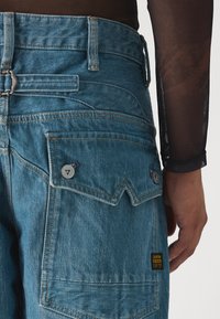 Jeans de ganga azul com um corte descontraído, costuras visíveis, dois bolsos traseiros com fechos de botão e uma etiqueta amarela de marca ao lado.