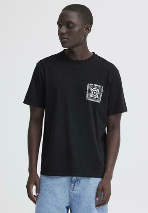 Noor mees, kes kannab musta T-särki valge "Sun Seekers" graafikaga rinnataskul, sellega on kombineeritud helesinised teksapüksid.