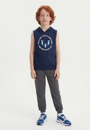 Παιδί με κόκκινα μαλλιά φορά αμάνικο μπλε σκούρο φούτερ με κουκούλα και το κείμενο «Ακολούθησε τα όνειρά σου Μην τα παρατάς ποτέ», γκρι φόρμα και μπλε αθλητικά παπούτσια.